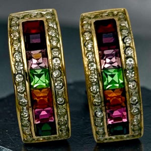 May include: A pair of gold-toned hoop earrings featuring a central vertical row of square-cut multicolored gemstones in red, purple, light pink, green, orange, and dark pink, bordered by two rows of small clear round crystals on each side.