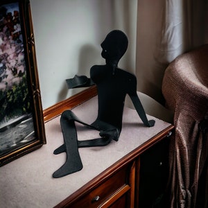 May include: A matte black metal sculpture of a stylized human figure in a seated position with legs extended and one arm raised. The figure is constructed from flat, bent metal sheets, featuring a smooth, dark finish and a minimalist silhouette.