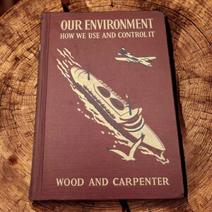 Puede incluir: Un libro de tapa dura color granate titulado Our Environment: How We Use and Control It por Wood y Carpenter. La portada presenta una ilustración color crema de un gran barco navegando, un avión volando y un ave en pleno vuelo.