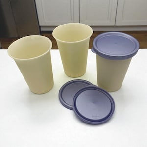 May include: Three beige plastic cups with a tapered design, accompanied by two dark purple lids. One cup has a lid on it. The cups are on a white surface, suggesting a kitchen or dining setting.
