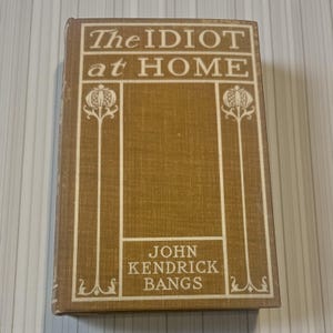 May include: A vintage book titled "The Idiot at Home" by John Kendrick Bangs. The book has a tan cover with white text and decorative floral elements. The cover has a textured appearance.
