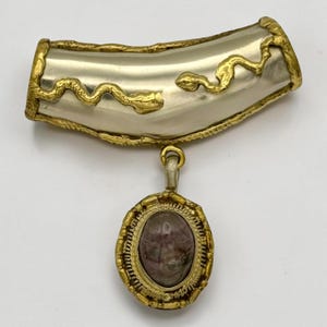 May include: A vintage brooch featuring a silver-tone curved bar with gold-tone snake detailing. A gold-tone chain connects to an oval pendant with a dark, speckled stone, framed by a gold-tone border. The brooch has an antique aesthetic.