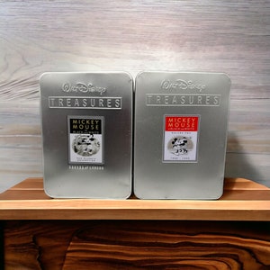 May include: Two silver metal boxes from the Walt Disney Treasures collection. The boxes feature the text "Treasures" and "Mickey Mouse" with black and white imagery. One box is labeled "Black and White" and the other "Volume Two".