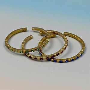 May include: Three gold-colored cuff bracelets. Each bracelet is adorned with small, colorful gemstones. One bracelet has green gems, another has red gems, and the third has blue gems. The bracelets are arranged on a white and blue gradient background.