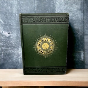 May include: A vintage book with a dark green cover. The cover features gold lettering that reads "GLEAMS OF GRACE" within a sunburst design. Decorative floral patterns border the top and bottom edges of the book.