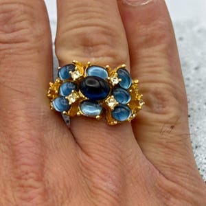 May include: A gold-toned ring featuring a cluster of oval-shaped blue cabochon stones and small clear accent stones. The central stone is a dark navy blue, surrounded by lighter blue stones arranged in a textured, organic metal setting.