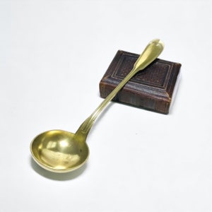 May include: A gold-toned metal ladle with a round bowl and a long, slender handle featuring a heart-shaped terminal. The ladle rests diagonally across a small, rectangular, dark brown leather-bound book with a textured, embossed cover design.