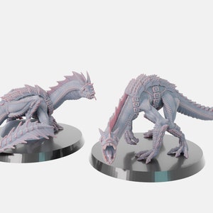 Puede incluir: Dos figuras en miniatura, grises y rosas, con forma de dragón, con colas y aletas espinosas. Las figuras están posadas sobre bases circulares negras.