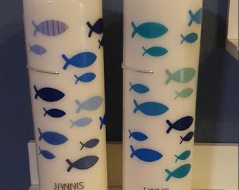 May include: Two white baptism candles with blue and teal fish designs. The candles have the names "Jannis" and "Linus" with the dates "10. April 2022" printed on them.