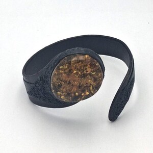 Könnte beinhalten: Schwarzes Armband mit strukturierter Band und einem großen, runden, bernsteinfarbenen Stein. Das Armband besteht aus einem dunklen Material und hat ein einzigartiges, organisches Design. Der Stein hat eine Mischung aus Braun- und Goldtönen.