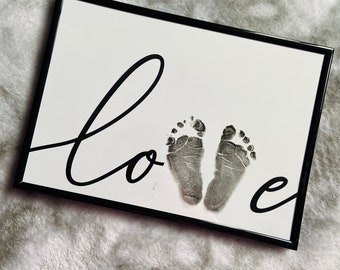 May include: A black framed print with the word "love" written in cursive and two baby footprints in the center.