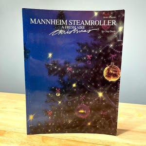 Op de afbeelding: De omslag van een muziekboek, "Mannheim Steamroller A Fresh Aire Christmas", toont een versierde kerstboom met ballen en lichtjes. De titel is wit, met de naam van de auteur, Chip Davis, bovenaan, tegen een donkerblauwe achtergrond.