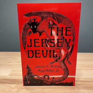 May include: A red book titled "The Jersey Devil" with a black illustration of a winged creature. The book's cover also features the names James F. McCloy and Ray Miller, Jr. The book is standing upright.