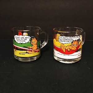 May include: Two glass mugs with Garfield cartoon characters. The mug on the left features Garfield on a seesaw with the text "I'm not one who rises to the occasion". The mug on the right features Garfield in a hammock with the text "It's not a pretty life but somebody has to live it".