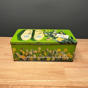 Könnte beinhalten: Eine rechteckige grüne Blechdose mit Deckel, die Abbildungen von Holzschuhen und Tulpen zeigt. Die Dose ist mit gelben und weißen Tulpen und dem Text "dutch country biscuits" verziert.