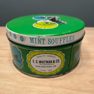 May include: A vintage green metal tin of "MINT SOUFFLES" with a white rim and a lid featuring a bicycle illustration. The tin has a yellow and green label that reads "E.G. Whitman & Co. Philadelphia, PA. Since 1854."