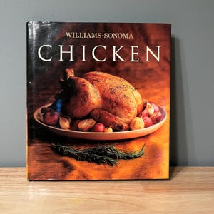 May include: A cookbook titled "CHICKEN" by Williams-Sonoma. The cover features a roasted chicken on a white plate with red potatoes and garlic. A sprig of rosemary is in the foreground. The background is a warm, brown color.