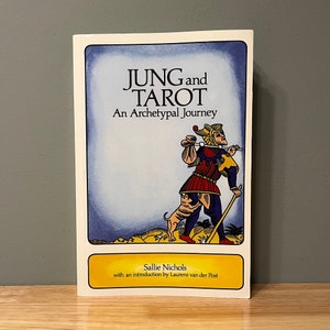 May include: A book titled "JUNG and TAROT: An Archetypal Journey" features a colorful illustration of a figure with a dog. The book's cover has a blue border and a yellow banner with the author's name, Sallie Nichols.