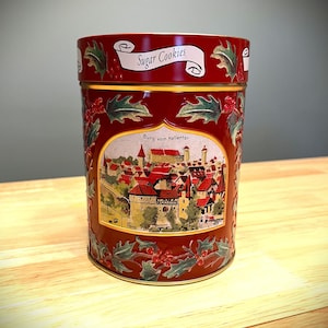 May include: A cylindrical, red metal cookie tin with a decorative design. The lid features the words "Sugar Cookies" on a white banner. The body of the tin displays a detailed illustration of a European village with red rooftops and the text "Burg vom Hallerfor".