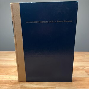May include: A hardcover book with a navy blue cover and a tan spine. The title "HOUSE & GARDEN'S COMPLETE GUIDE TO Interior Decoration" is printed in gold lettering. The book is on a wooden surface.