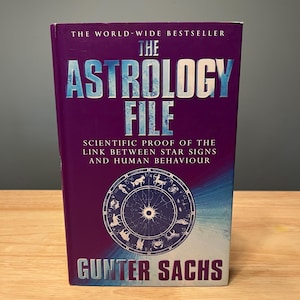 May include: A hardcover book titled The Astrology File by Gunter Sachs with a deep purple cover. The front features white text, a circular zodiac wheel graphic with twelve star signs, and the subtitle Scientific Proof of the Link Between Star Signs and Human Behaviour.