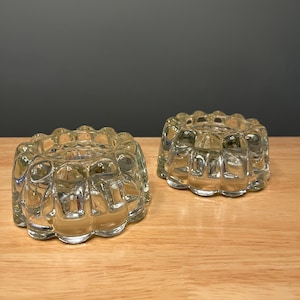 Puede incluir: Dos candelabros de vidrio transparente con un diseño acanalado y redondeado. Los candelabros están hechos de vidrio transparente y se asientan sobre una superficie de madera. Los candelabros son redondos y tienen un borde festoneado.