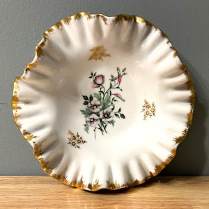 Op de afbeelding: Een witte porseleinen kom met een gegolfde rand en gouden rand. De kom heeft een bloemenmotief met roze en witte bloemen en groene bladeren.