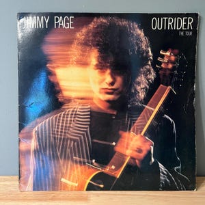 May include: A vintage vinyl record album cover featuring Jimmy Page, a musician, holding a guitar. The album cover is a dark background with a blurred image of Jimmy Page in a striped jacket. The album title is "Outrider: The Tour".