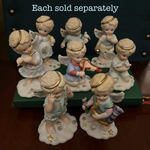 Peut inclure: Ensemble de huit figurines d'anges en porcelaine, chacune avec une pose différente et tenant un objet différent. Les anges sont tous vêtus de blanc et de bleu avec des accents dorés. Chaque ange est debout sur une base en forme de nuage avec des étoiles dorées. Le texte "Each sold separately" est visible en haut de l'image.