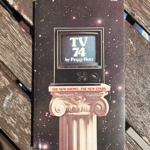 May include: A vintage book cover with a black and white illustration of a television set. The television screen displays the text "TV 74 by Peggy Herz". The book cover has a dark background with white stars and the text "THE NEW SHOWS...THE NEW STARS" in red.