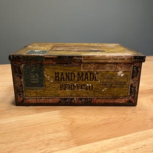 Puede incluir: Una caja de lata rectangular antigua con un aspecto envejecido y desgastado. La caja es principalmente marrón y dorada, con el texto "HAND MADE PERFECTO". La caja tiene una tapa con bisagras y parece ser de metal. La caja está sobre una superficie de madera.
