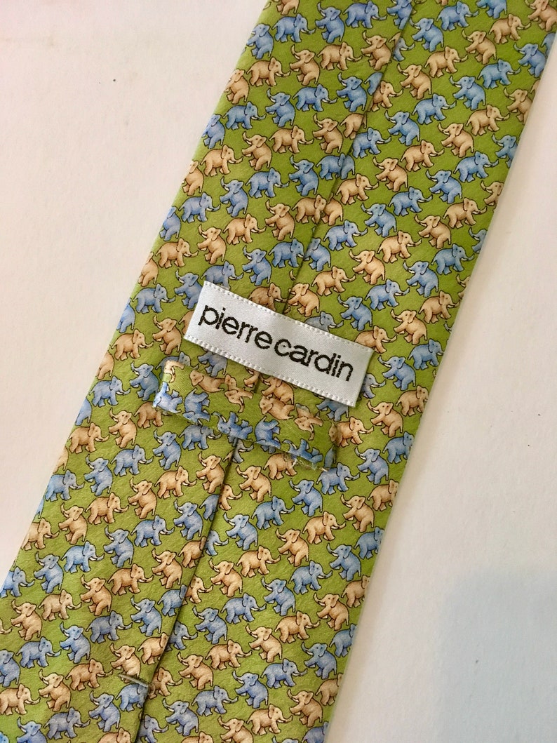 K&ouml;nnte beinhalten: Eine gr&uuml;ne Seidenkrawatte mit einem sich wiederholenden Muster aus hellblauen und beigen Elefanten. Die Krawatte hat ein wei&szlig;es Etikett mit dem Text "pierre cardin".
