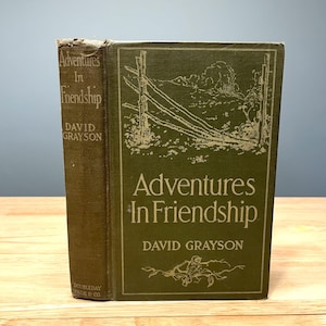 May include: A vintage hardcover book titled "Adventures In Friendship" by David Grayson. The book has a green cover with gold lettering and a faded illustration of a fence and landscape. The spine shows the title and author.