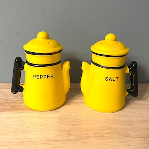 May include: Yellow ceramic salt and pepper shakers shaped like coffee pots with black handles. The shakers have black lettering that reads "SALT" and "PEPPER".
