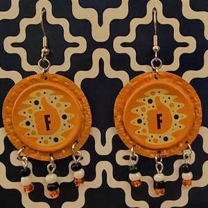 May include: A pair of orange bottle cap earrings. Each earring features a thumbs-up design with the letter "F" in the center. Dangling from each earring are small beaded accents in black, white, and orange.