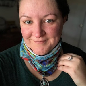 Pode incluir: Uma mulher usando um cachecol de pescoço estampado em azul-turquesa, azul e vermelho com um colar de pingente de prata. O pingente é oval com um design de árvore e pedras coloridas.