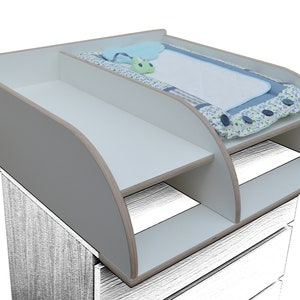 Op de afbeelding: Een wit en grijs aankleedtafel met twee planken en een gebogen ontwerp. De bovenste plank bevat een blauwe aankleedkussen met een bloemenrand en een klein speeltje. De tafel is ontworpen voor babyverzorging.