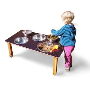 Könnte beinhalten: Ein Holz-Aktivitätstisch mit vier Edelstahlschalen und einer gelben Schaufel. Der Tisch hat Holzbeine und Rollen. Ein Kind spielt mit dem Tisch.