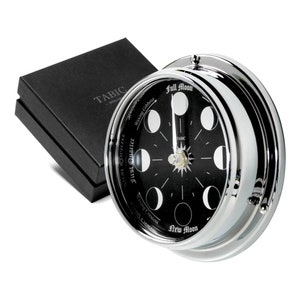 May include: A chrome-finished clock with a black face displaying the phases of the moon. The clock face features the text "Full Moon" and "New Moon". A black box with the text "TABIC" is in the background.