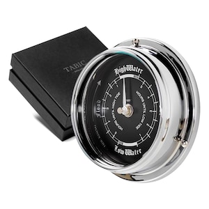 Può includere: Un orologio a marea cromato con un quadrante nero. L'orologio ha un ago argentato e segni per le ore fino all'alta marea e le ore fino alla bassa marea. L'orologio indica "Alta marea" e "Bassa marea" nella parte superiore e inferiore del quadrante.