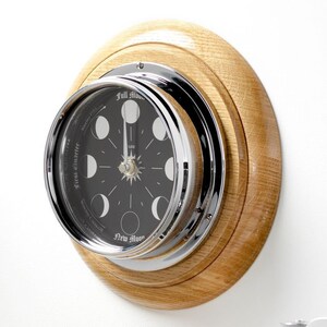May include: A round, black and white moon phase clock with a chrome bezel and oak wood frame. The clock face shows the phases of the moon, with the words "Full Moon", "First Quarter", "Waning Crescent", "New Moon", and "Waxing Crescent" printed around the edge.
