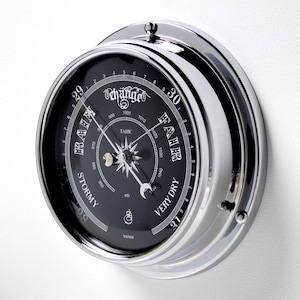 May include: A chrome-plated barometer with a black face and a white dial. The dial has markings for inches and millibars, and the face has a weather forecast scale with markings for "Very Dry", "Fair", "Stormy", and "Hurricane".