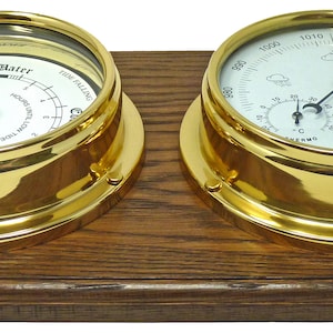 May include: A brass nautical barometer and hygrometer set mounted on a wooden base. The barometer has a white face with black markings and a needle pointing to the current barometric pressure. The hygrometer has a white face with black markings and a needle pointing to the current humidity level. The set is a decorative and functional piece for any home or office.