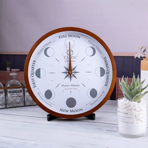 May include: A wooden moon phase clock with a brown frame and white face. The clock features a compass-like design with a needle pointing to the current moon phase. The phases are labeled: New Moon, Waxing Crescent, First Quarter, Waxing Gibbous, Full Moon, Waning Gibbous, Third Quarter, and Waning Crescent. The clock also includes the text "TABIC Pretece" and "Moon-Master Great British Design."
