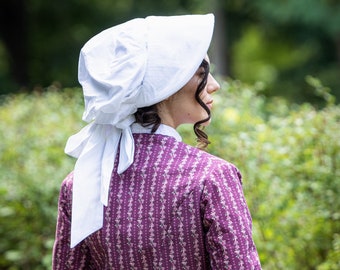 May include: A woman wearing a purple and white floral dress with a white bonnet. The dress has a white sash tied at the waist.