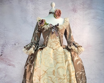 May include: A historical-style gown in shades of brown and cream, displayed on a mannequin. The bodice and sleeves are brown with ornate silver embroidery, while the skirt is cream-colored with a gold floral pattern. The gown is adorned with lace and floral accents.