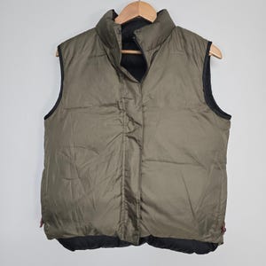 May include: Olive green puffer vest with a black interior and trim. The vest has a high collar and a full-length zip closure. The vest is hanging on a wooden hanger.