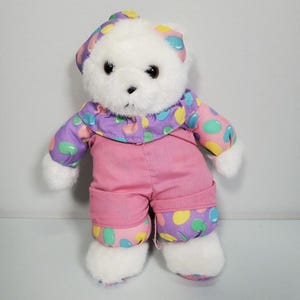 May include: A white teddy bear wearing a pink outfit with a purple jacket and hat. The jacket and hat are decorated with colourful polka dots. The bear has black eyes and a black nose.