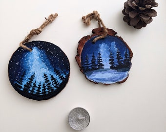 May include: Two round wood slices with painted scenes of a forest at night. One slice features a blue sky with stars and a white mountain peak. The other slice features a blue sky with clouds and a dark blue lake. Both slices have a brown twine hanger.