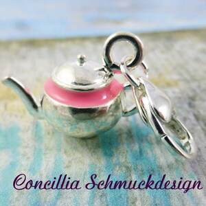 Könnte beinhalten: Ein silberner Teekannen-Anhänger mit einem rosa Band. Der Anhänger hat eine kleine Schlaufe und einen Verschluss zum Befestigen an einem Armband oder einer Halskette. Der Text "Concillia Schmuckdesign" befindet sich unten.